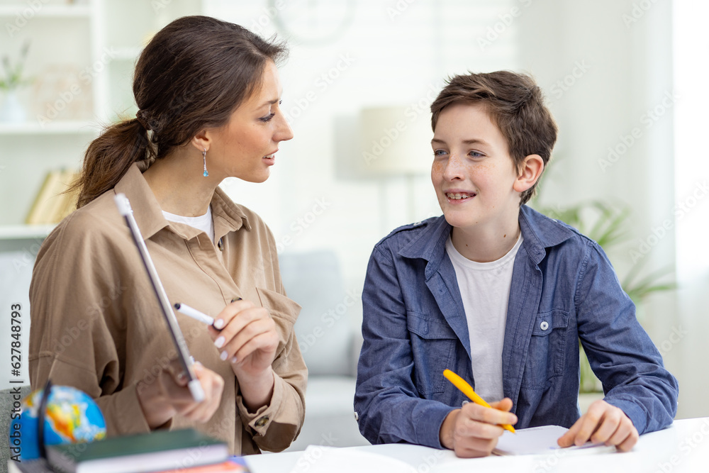 Home education, woman teaches boy at home sitting at table in living room, mother explains educational material to son doing homework together. Home education, woman teaches boy at home sitting at table in living room, mother explains educational material to son doing homework together.