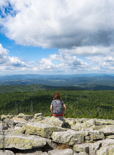 brown-haired female hiker with backpack sitting on rock overlooking forest on blue sky day