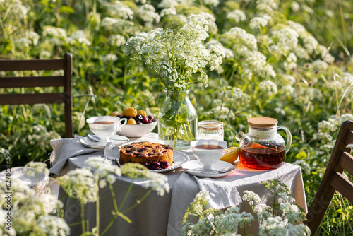Fancy white porcelain set for herbal tea or coffee and homemade pie on wooden table in the garden. Summer outdoor party arrangement, romantic date. White flowers on background and in a glass vase