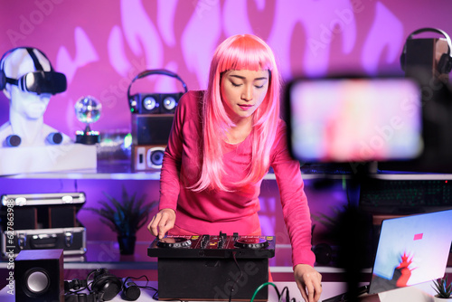 Smiling woman mixing techno music at professional mixer console having fun while performing song at night in club. Artist with pink hair recording performace with camera posting online for subscribers photo