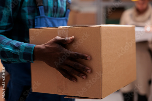 African american warehouse worker man hands holding cardboard package. Storehouse employee arms carrying carton container with adhesive tape close up, working in stockroom