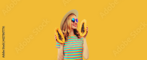 Summer portrait of happy cheerful young woman having fun with fresh papaya fruits wearing straw hat, sunglasses on yellow background