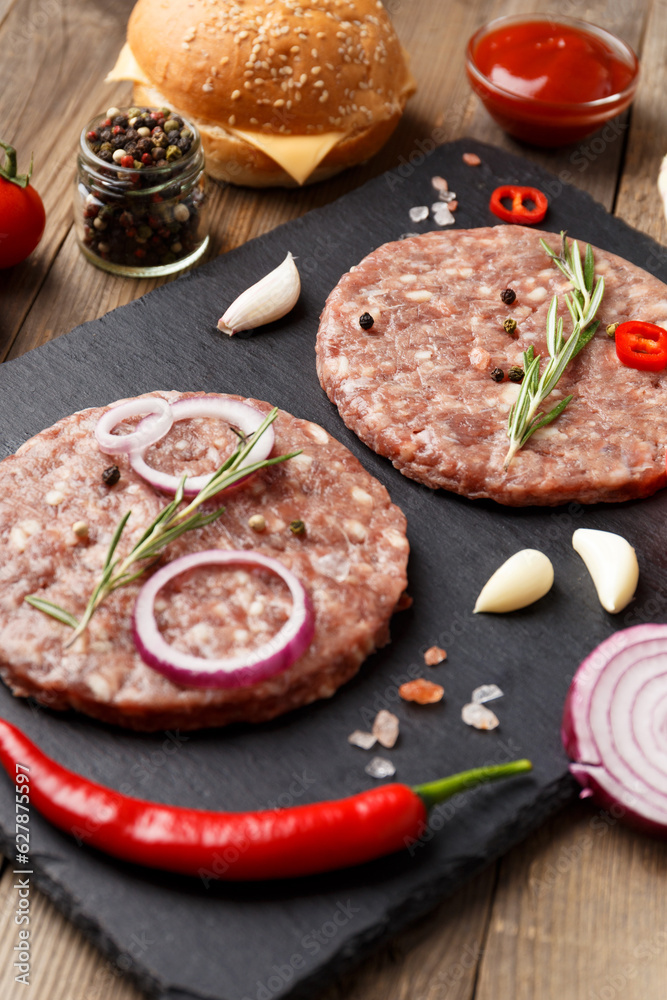 Raw beef hamburger patties on stone plate, vegetables and spices, wooden background.