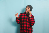 Shocked Young Asian man with a beanie hat and a red plaid flannel shirt is pointing his finger at copyspace while talking on his mobile phone, isolated on a blue background