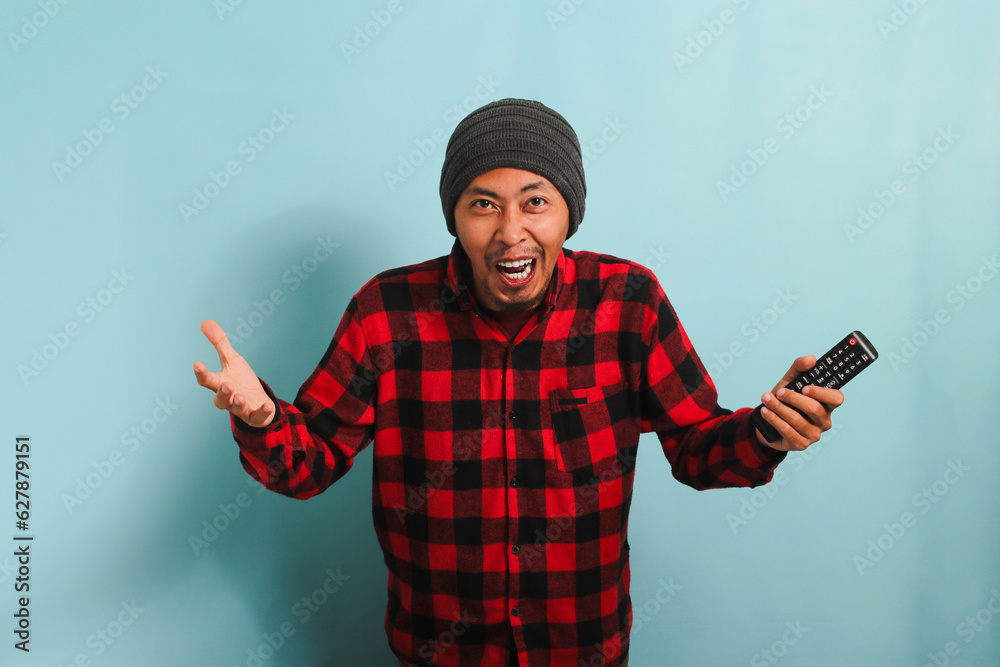 Excited young Asian man with a beanie hat and red plaid flannel shirt watching TV and cheering for a sports competition with laughter and a sense of victory, isolated on a blue background