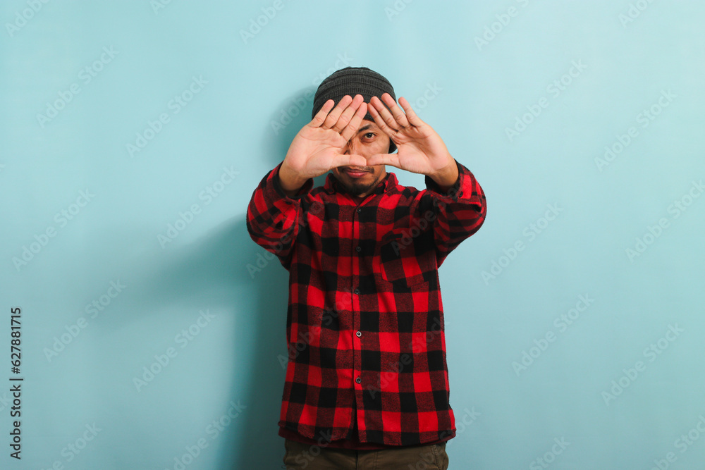 Young Asian man with beanie hat and red plaid flannel shirt making a triangle gesture with palms, symbolizing the environment protection, recycling, and reusing concept, isolated on a blue background