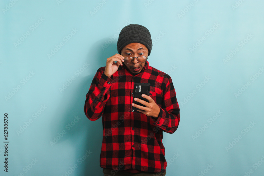 Surprised young Asian man with beanie hat and red plaid flannel shirt, his glasses raised, receives great news or special offer while using his smartphone and online shop, isolated on blue background Surprised young Asian man with beanie hat and red plaid flannel shirt, his glasses raised, receives great news or special offer while using his smartphone and online shop, isolated on blue background