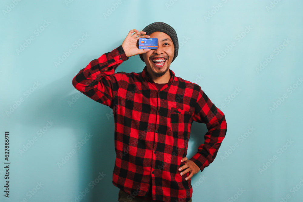 Joyful young Asian man with a beanie hat and red plaid flannel shirt is playfully covering one eye with a credit card while standing against a blue background