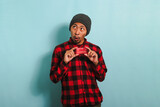 Young Asian man with a beanie hat and red plaid flannel shirt is holding bank credit cards in his hands, with a surprised expression looking at copy space while standing against a blue background.
