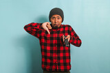 Displeased young Asian man with a beanie hat and red plaid flannel shirt giving a thumbs-down to a bad TV show, isolated on a blue background