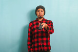 An annoyed young Asian man with a beanie hat and red plaid flannel shirt is switching TV channels using the TV remote, feeling upset and bored, isolated on a blue background.