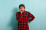 Unhappy Young Asian man with beanie hat and red plaid flannel shirt holding his cheek with his hand and showing a painful expression due to toothache or dental illness, isolated on a blue background