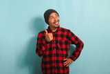 A confident young Asian man with a beanie hat and a red plaid flannel shirt is giving a thumbs-up while looking at right copy space, expressing a positive review. He is isolated on a blue background