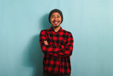 A confident young Asian man with a beanie hat and a red plaid flannel shirt is looking at the camera with his arms crossed. He is isolated on a blue background