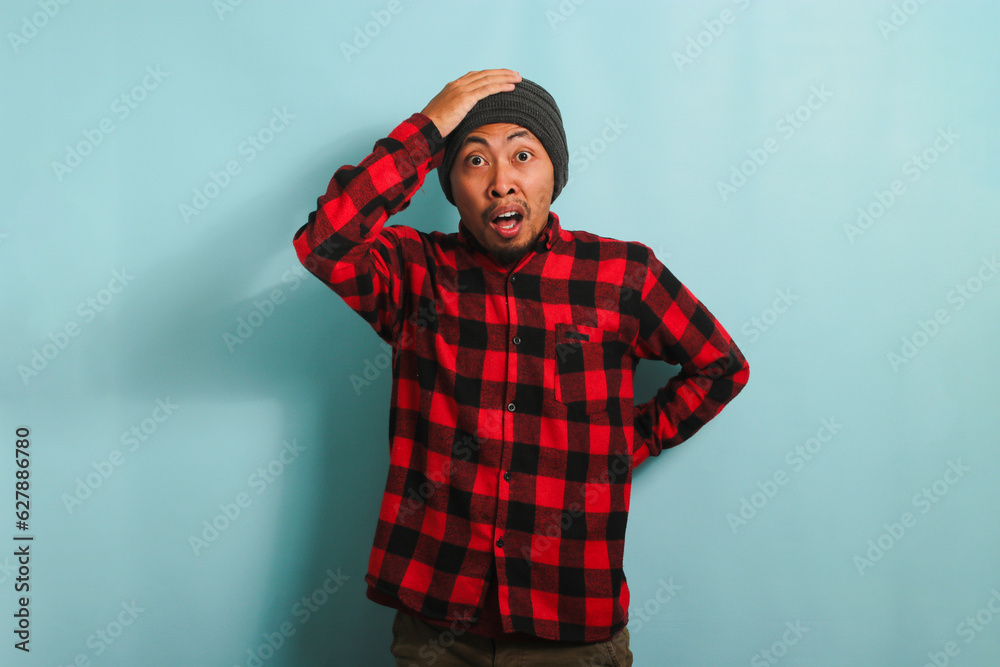 Scared Young Asian man with a beanie hat and red plaid flannel shirt has an open mouth, grabs his head, and looks at the camera reacting to something horrible, isolated on a blue background