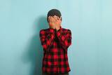 An ashamed and embarrassed young Asian man with a beanie hat and a red plaid flannel shirt facepalms, covering his face with his hand while standing against a blue background