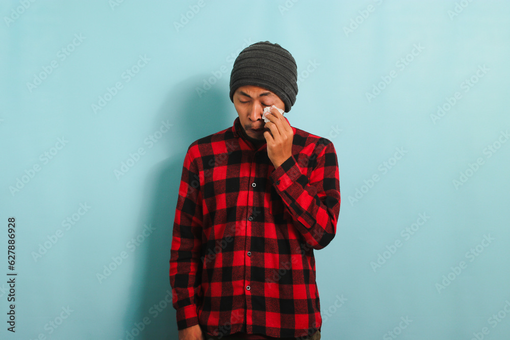 A crying young Asian man with a beanie hat and a red plaid flannel shirt wipes tears with a tissue while standing against a blue background A crying young Asian man with a beanie hat and a red plaid flannel shirt wipes tears with a tissue while standing against a blue background