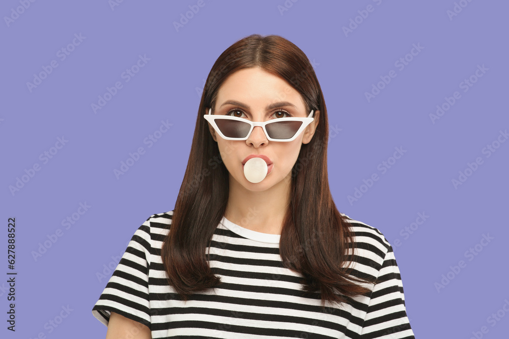 Beautiful woman in sunglasses blowing bubble gum on light purple background