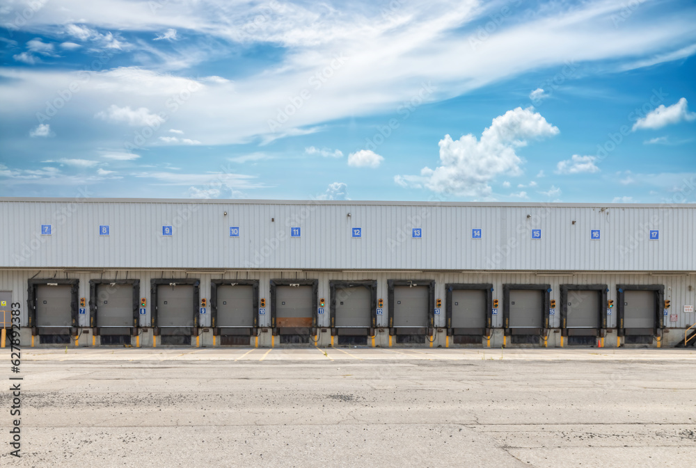 High bay warehouse facade showing long row of empty numbered truck loading bays, rubber door seals, target lights, empty asphalt parking lot in foreground, blue sky with clouds, nobody