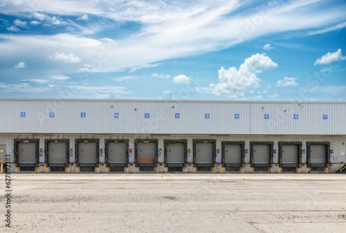 High bay warehouse facade showing long row of empty numbered truck loading bays  rubber door seals  target lights  empty asphalt parking lot in foreground  blue sky with clouds  nobody