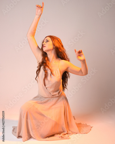 Full length portrait of beautiful brunette female model wearing dress with golden back light lighting. Kneeling pose with gestural arm pose. isolated on studio background.