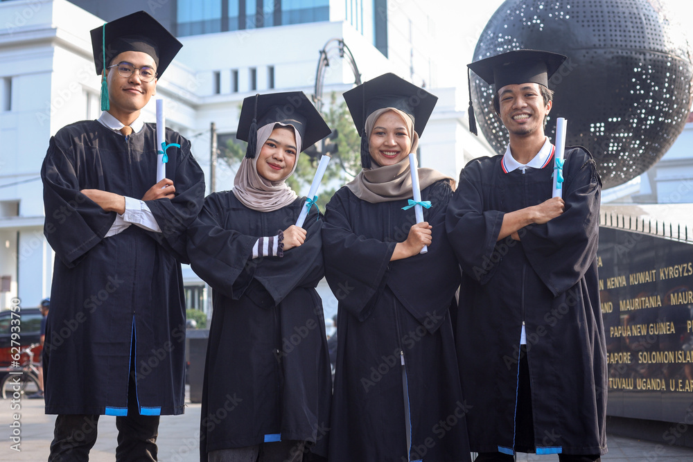 Obraz premium Portrait of four college graduates in graduation gowns standing close to each other, holding certificate of diploma and looking at camera smiling.