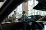 Remote view from inside of car through windshield to handsome smiling car dealer talking with cheerful male in showroom and shaking hand. Cheerful two people standing in dealership and talking.