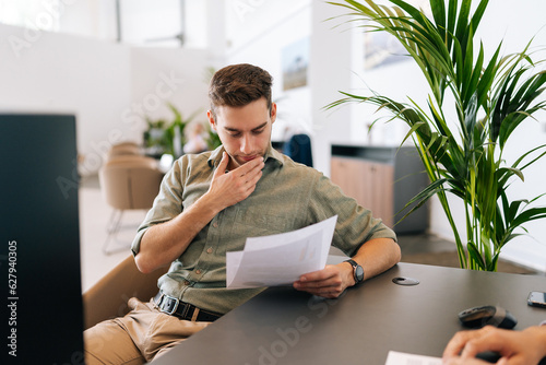 Portrait of focused male client reading contract for real estate property purchase. Thoughtful man reading terms of conditions of document, making decision about house purchase or financial investment photo