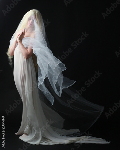 Full length portrait of beautiful blonde woman wearing white gown dress with flowing ghostly veiled fabric, isolated on dar studio background.