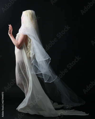 Full length portrait of beautiful blonde woman wearing white gown dress with flowing ghostly veiled fabric, isolated on dar studio background.