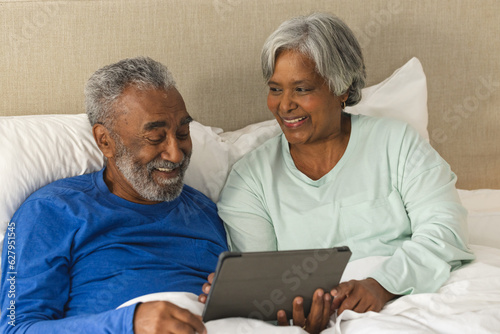 Happy senior biracial couple lying in bed using tablet at home