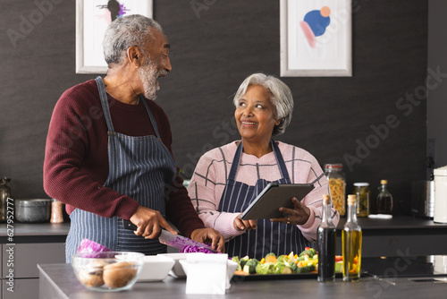 Happy senior biracial couple in aprons using tablet preparing vegetables in kitchen at home