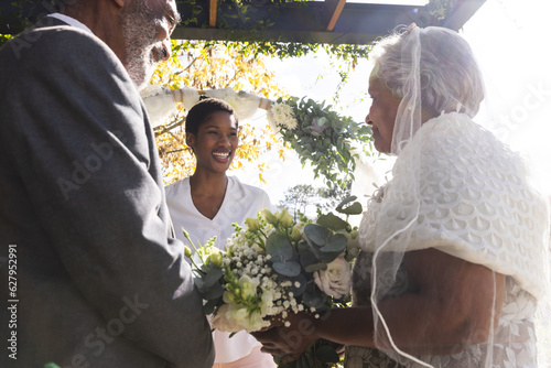 Happy biracial female marriage officiant with senior couple during wedding ceremony in sunny garden