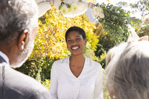 Happy biracial female marriage officiant and senior couple during wedding in sunny garden