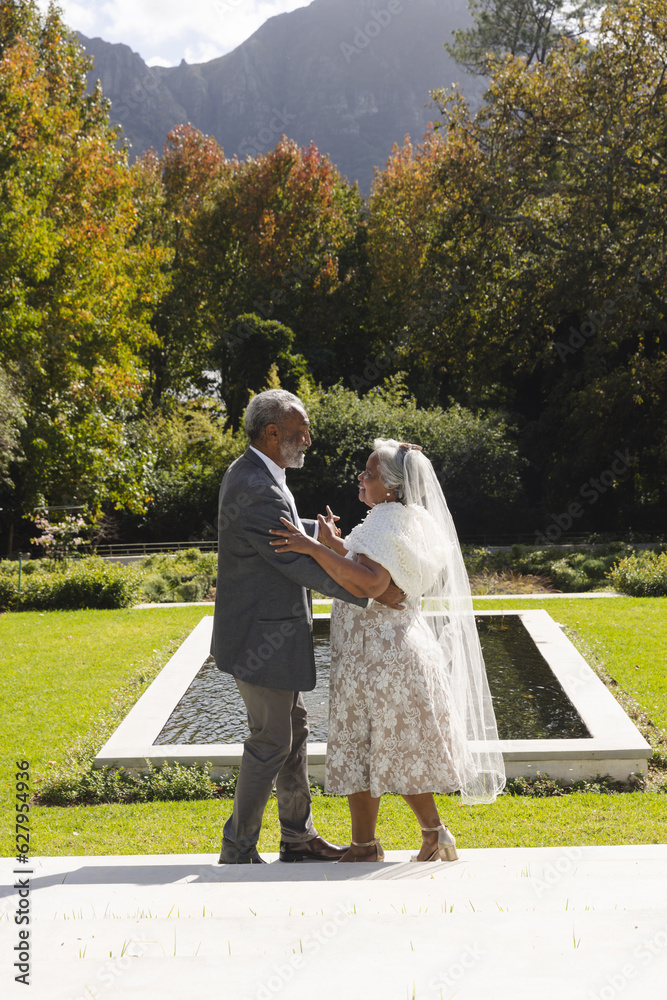 Fototapeta premium Happy senior biracial bride and groom dancing in garden at sunny wedding ceremony, copy space