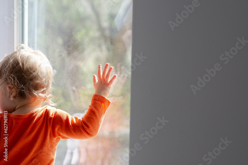 baby girl standing in front of the window and touch glass at sunny day, having fun with the sunbeams, natural dirty home glass