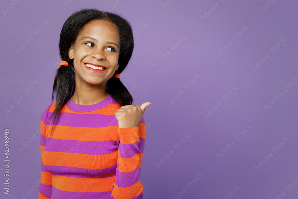 Little kid teen girl of African American ethnicity 15-16 year old wear striped orange sweatshirt point thumb finger aside back on area isolated on plain purple background. Childhood lifestyle concept. Little kid teen girl of African American ethnicity 15-16 year old wear striped orange sweatshirt point thumb finger aside back on area isolated on plain purple background. Childhood lifestyle concept.