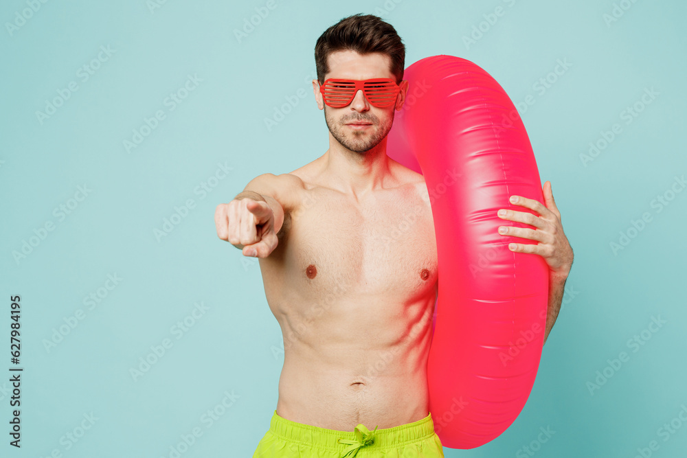 Young man wear green shorts swimsuit relax near hotel pool hold inflatable rubber ring point index finger camera on you isolated on plain blue cyan background Summer vacation sea rest sun tan concept