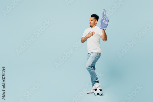 Full body young man fan in t-shirt foam 1 fan glove finger up sing national country anthem cheer up support football sport team hold soccer ball watch tv live stream isolated on plain blue background. photo
