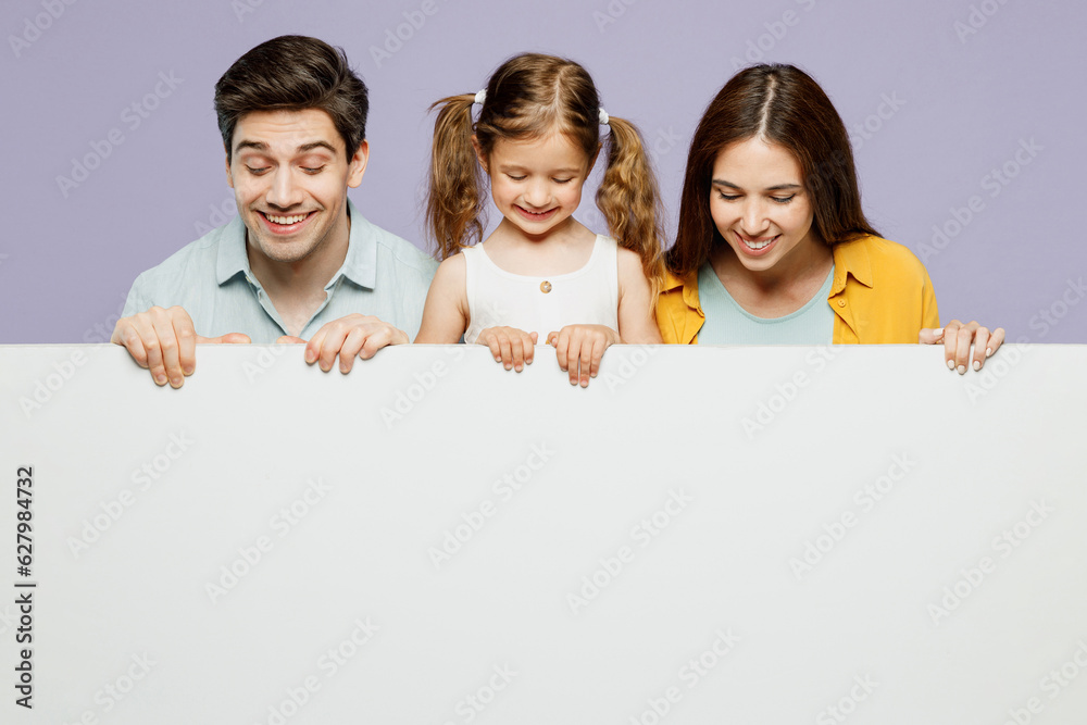 Young parents mom dad with child kid daughter girl 6 years old wearing blue yellow casual clothes hold look at big white empty poster billboard isolated on plain purple background. Family day concept. Young parents mom dad with child kid daughter girl 6 years old wearing blue yellow casual clothes hold look at big white empty poster billboard isolated on plain purple background. Family day concept.