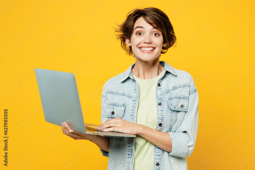 Young surprised shocked happy happy IT woman wear green t-shirt denim shirt casual clothes hold use work on laptop pc computer isolated on plain yellow background studio portrait. Lifestyle concept. Young surprised shocked happy happy IT woman wear green t-shirt denim shirt casual clothes hold use work on laptop pc computer isolated on plain yellow background studio portrait. Lifestyle concept.