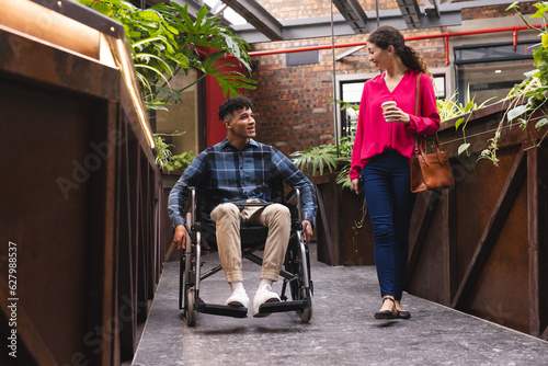 Happy diverse female and male colleague in wheelchair talking in office corridor photo