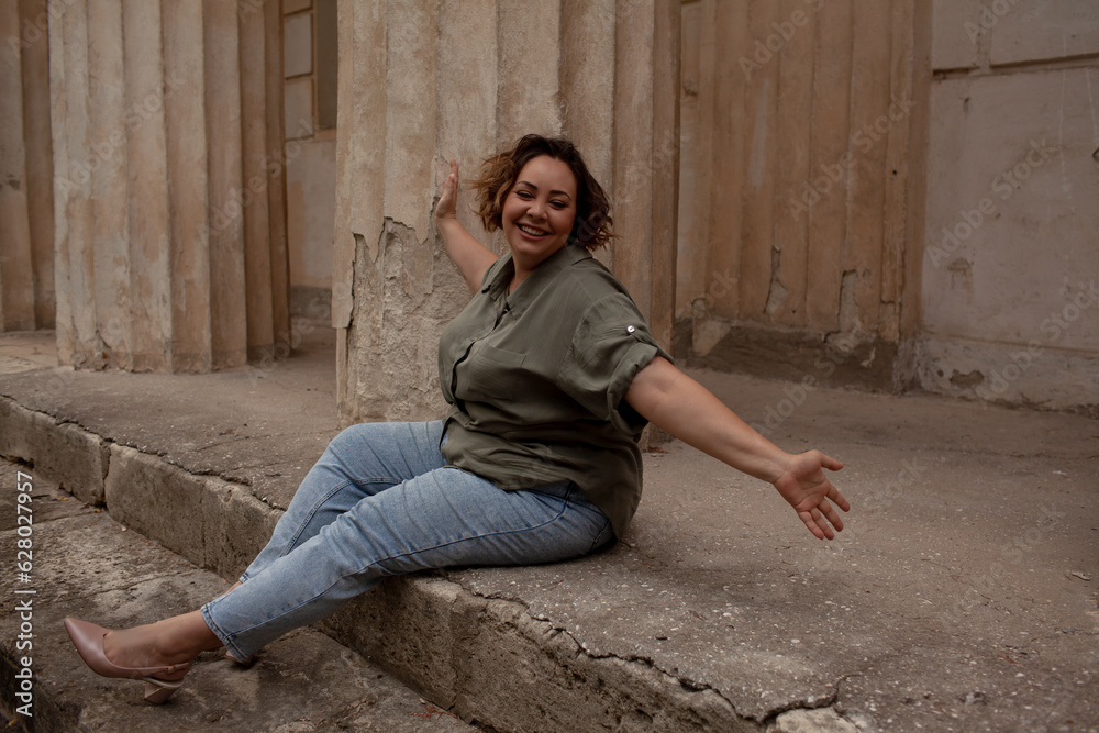 A beautiful girl is wearing a khaki t-shirt and jeans, the overweight woman posing against stone biege wall. The fashion outfit for plus-size woman for the city, minimalizm urban style clothes