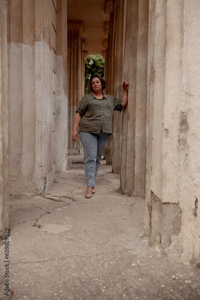 A beautiful girl is wearing a khaki t-shirt and jeans, the overweight woman posing against stone biege wall. The fashion outfit for plus-size woman for the city, minimalizm urban style clothes