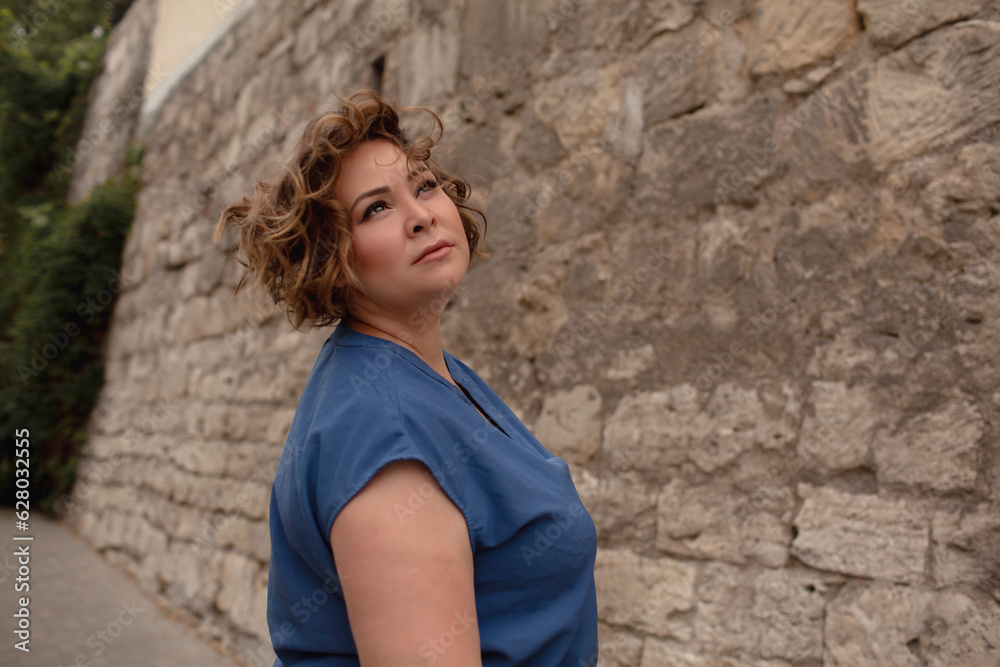 portrait of a beautiful girl is wearing a blue t-shirt, the overweight woman posing against stone biege wall. The fashion outfit for plus-size woman for the city, minimalizm urban style clothes portrait of a beautiful girl is wearing a blue t-shirt, the overweight woman posing against stone biege wall. The fashion outfit for plus-size woman for the city, minimalizm urban style clothes