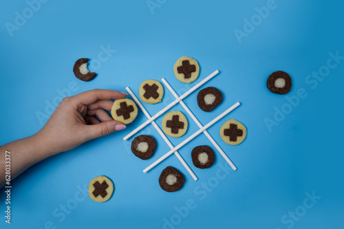 Tic tac toe game with freshly baked cookies and a woman's hand reaches for one of the cookies on blue background top view