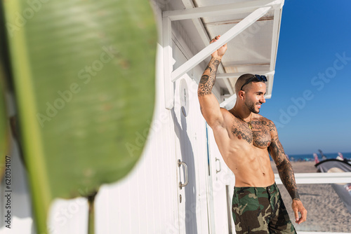 Chico joven musculoso y tatuado posando sin camiseta y en bañador frente a chiringuito de playa photo