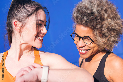 close up portrait of two multiethnic friends using a smart watch while practicing sport, concept of sports technology and active lifestyle, copy space for text