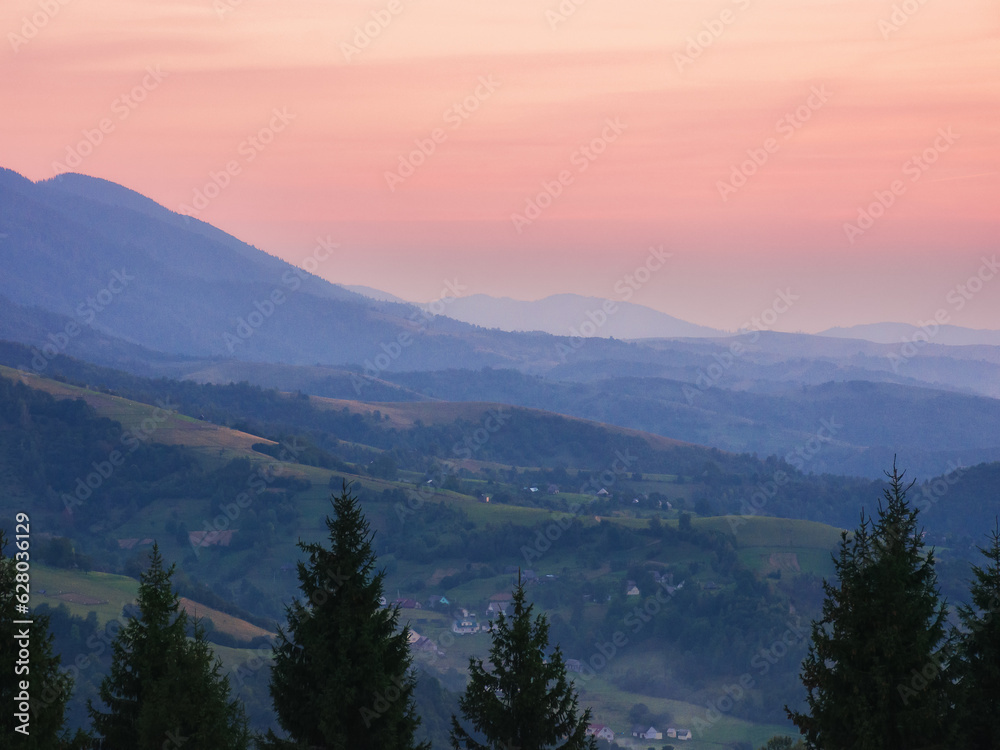 Obraz premium sunset in carpathian mountains. distant rural valley with rolling hills between forested slopes in hazy atmosphere. pink sky