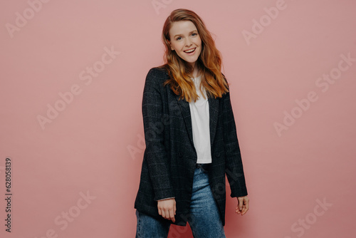 Stylish young woman in plaid blazer and jeans, posing carefree and expressing positive emotions in studio, isolated on pink wall background. Happy people concept.